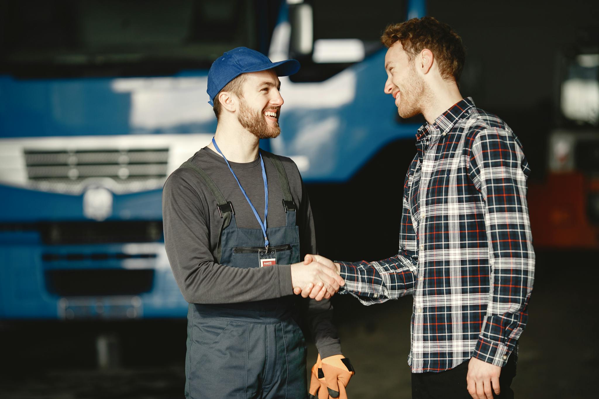 Mechanic in uniform and customer in plaid shirt shaking hands in front of a blue truck inside a garage.